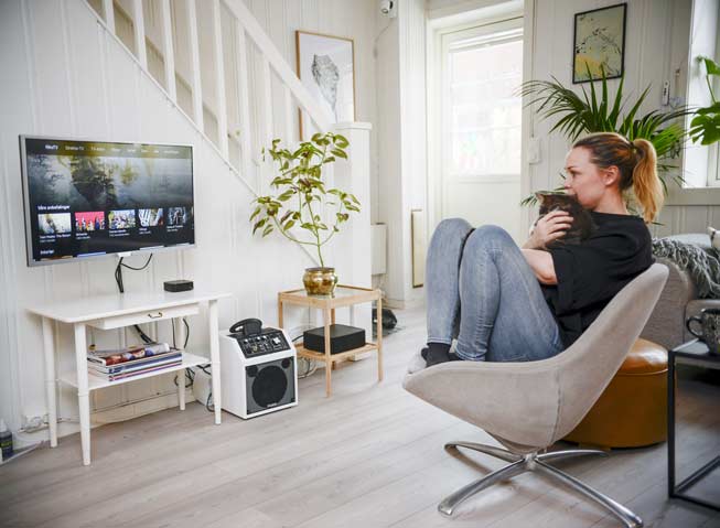 Kvinne sittende i stol i stue, med katt i armkroken, mens hun velger program på TVen via RiksTV