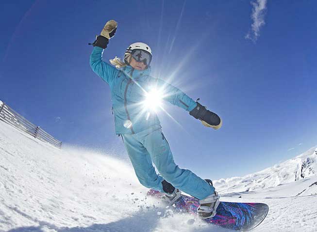 Kristin Krohn Devold susende ned en skibakke på snowboard, i solskinn, under en gnistrende blå himmel.