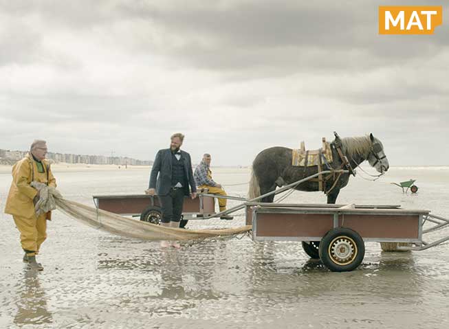 To fiskere og en programmleder i dress står på en bred sandstrand i Flandern med to hester, kjerrer og garn.