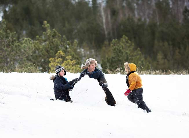 Far sammen med to barn i snøkledt landskap, bygger snømann