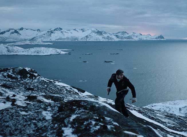 Jan Baalsrud løpende opp en fjellside i spektakulært Nord-Norsk landskap, på kveldstid, med en pistol i hånden.