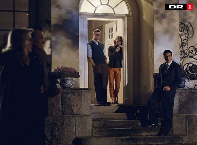 To personer står i en dørkarm, til høyre Tuva Novotny i rollen som Puck. Det er mørkt ute, folk går forbi, de to ser bekymrede ut. På trappa står en tredje mann i dress.