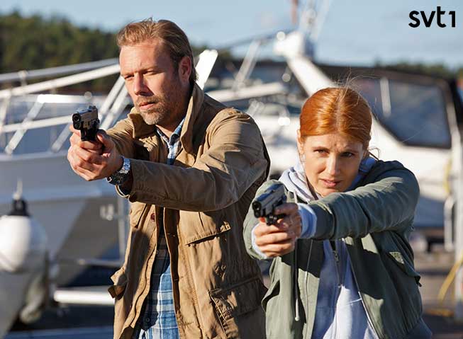 To politifolk - en mann og en kvinne - side om side i en småbåthavn, med pistoler pekende framover.